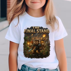 以下が含まれることがあります： 「THE FINAL STAND」の文字とカラフルなグラフィックがプリントされた白いTシャツ。グラフィックは、森の中でピクセル化されたキャラクターを描いています。人物は青いジーンズのショートパンツとピンクの野球帽を着用しています。