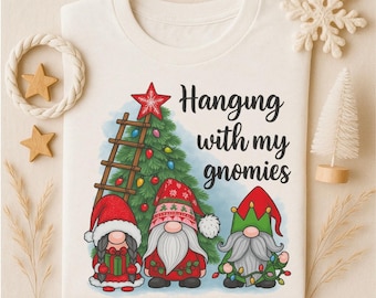 May include: Cream-colored t-shirt featuring a Christmas gnome design. The graphic includes three gnomes, a Christmas tree, a ladder, and the phrase "Hanging with my gnomies." The gnomes wear red and green hats and hold Christmas lights.