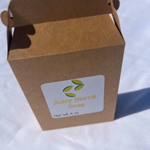 May include: A brown cardboard box with a decorative top and a white label. The label reads "Juicy Neroli Soap" with a leaf design and "Net Wt. 6 oz."