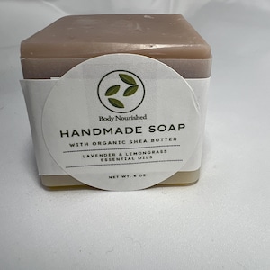 May include: A square bar of handmade soap with a light brown top and a white label. The label reads "Body Nourished Handmade Soap with Organic Shea Butter Lavender & Lemongrass Essential Oils." The net weight is 227 g.