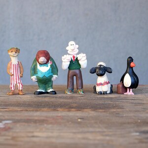 May include: A set of six plastic figurines, including a penguin, a sheep, a man with a green sweater, a mole wearing a tuxedo, and an otter wearing a red and white striped shirt.