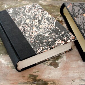 Op de afbeelding: Twee zwarte hardcover boeken met marmerpapier omslagen. De boeken zijn gesloten en liggen op een houten oppervlak.