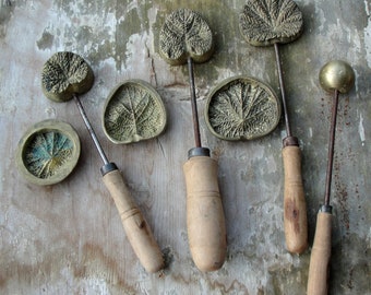 May include: Five antique brass leaf-shaped cookie cutters with wooden handles. The cutters are arranged on a light brown wooden surface.