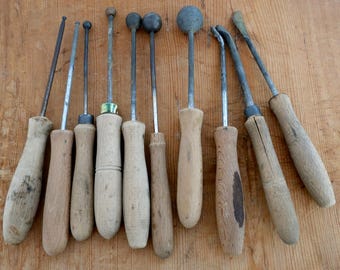 May include: A collection of antique metal tools with wooden handles, including various shapes like spheres and flat blades. The tools are arranged on a light brown wooden surface, showcasing their aged appearance and craftsmanship.