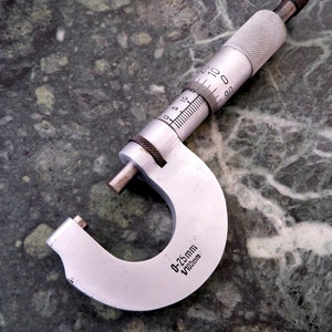 以下が含まれることがあります： 黒いノブと測定スケールが付いた銀色の金属製ノギス。ノギスには「0-25mm 1/1000mm」と表示されています。