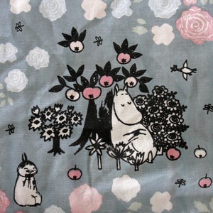 Könnte beinhalten: Ein hellblauer Stoff mit einer skurrilen Illustration eines Mumin-Charakters, schwarzen Bäumen, Blumen und rosa Äpfeln. Das Design ist mit weißen und rosa Rosen und kleinen schwarzen Blättern durchsetzt.