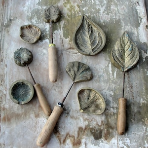 May include: A set of seven metal leaf and flower shaped cookie cutters with wooden handles. The cutters are arranged on a rustic wooden surface.