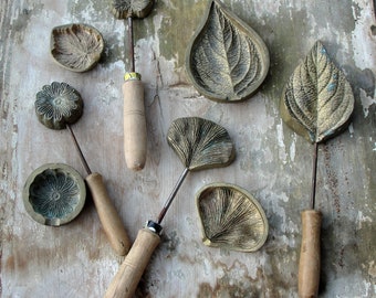May include: A set of seven metal leaf and flower shaped cookie cutters with wooden handles. The cutters are arranged on a rustic wooden surface.