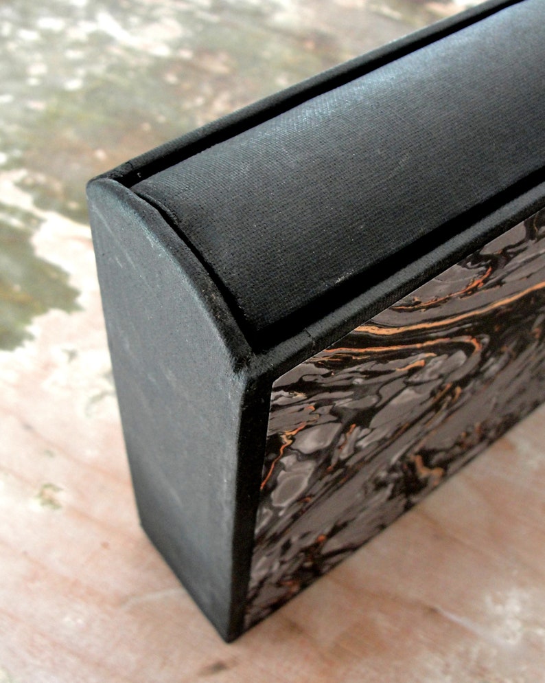 K&ouml;nnte beinhalten: Eine schwarze Box mit einem Marmormuster an der Seite. Die Oberseite der Box ist mit schwarzem Stoff bedeckt.
