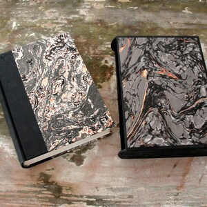 K&ouml;nnte beinhalten: Zwei schwarze und graue marmorierte Hardcover-B&uuml;cher mit schwarzen R&uuml;ckseiten. Die B&uuml;cher haben ein marmoriertes Muster mit schwarzen, grauen und wei&szlig;en Wirbeln. Die B&uuml;cher liegen auf einer Holzoberfl&auml;che.