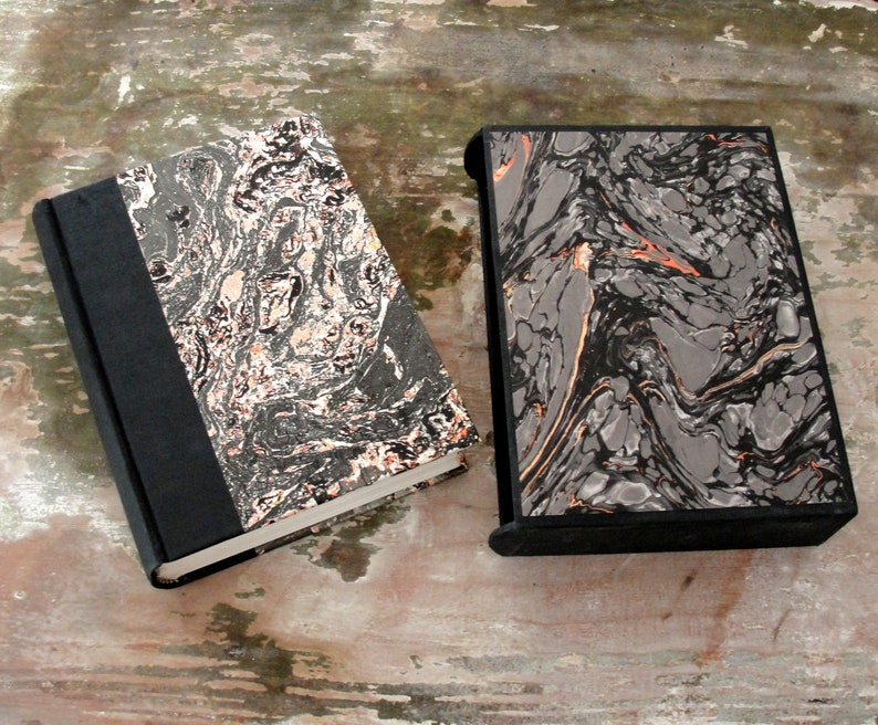 K&ouml;nnte beinhalten: Zwei schwarze und graue marmorierte Hardcover-B&uuml;cher mit schwarzen R&uuml;ckseiten. Die B&uuml;cher haben ein marmoriertes Muster mit schwarzen, grauen und wei&szlig;en Wirbeln. Die B&uuml;cher liegen auf einer Holzoberfl&auml;che.