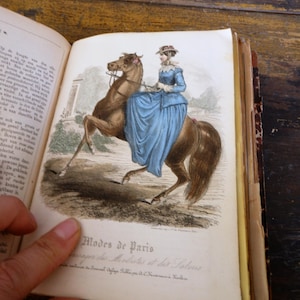 May include: An open antique book displays a color illustration of a woman in a blue dress riding a brown horse. The text "Modes de Paris" is visible below the illustration. The book's pages are aged and yellowed.