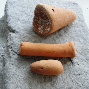 May include: Three terracotta pottery pieces on a gray stone surface. The pieces include a cone-shaped piece, a long cylinder, and a rounded oval.