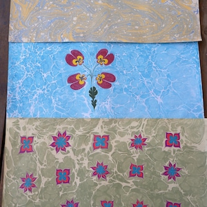 May include: Three sheets of decorative paper with marbled patterns. The top sheet has a light blue and yellow swirl design. The middle sheet is blue with a floral design. The bottom sheet is green with a floral pattern.
