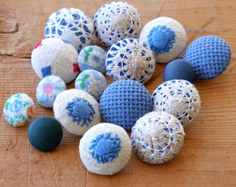 Puede incluir: Una colección de 15 botones vintage en varios tonos de azul, blanco y crema. Los botones están hechos de tela y tienen diferentes texturas, incluyendo crochet, bordado y tejido. Algunos botones tienen patrones florales.
