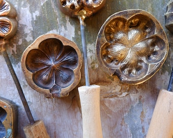 May include: Close-up of several antique brass flower-shaped waffle irons. Each iron has a detailed floral design on the cooking surface, with wooden handles and metal stems. The irons are arranged on a weathered wooden surface.