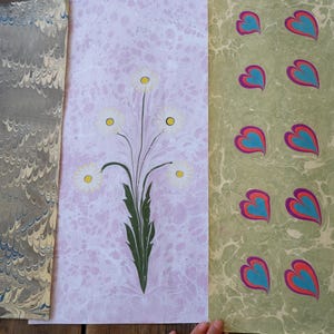 Puede incluir: Tres hojas de papel decorativo con patrones únicos. Una hoja tiene un diseño veteado gris y blanco, otra presenta un motivo floral con margaritas blancas, y la tercera tiene un patrón de corazones repetidos en turquesa y rojo.