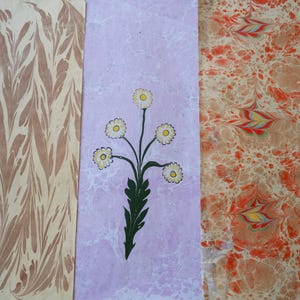 May include: Three rectangular pieces of paper with decorative patterns. The left piece has a cream and brown marbled design. The center piece is lavender with a floral illustration. The right piece has a peach and red marbled design.