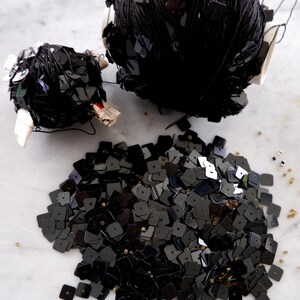 May include: Two spools of black square sequins, one is wrapped in black thread and the other is loose. The loose sequins are scattered on a white marble surface.