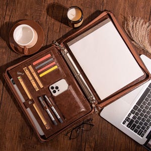 May include: A brown leather portfolio with a zipper, open to reveal a notepad, card slots, and a phone pocket. The portfolio also holds pens, a ruler, and a tablet. A cup of coffee, a candle, and a laptop are nearby.