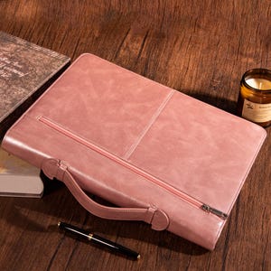 May include: A dusky pink leather portfolio with a handle and zip closure. The portfolio is on a wooden surface, with a pen and a lit candle in the background. The portfolio has a stitched design.