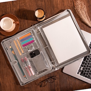 May include: A silver zippered portfolio with multiple compartments, including slots for pens, a phone, and colorful sticky notes. The portfolio is open, revealing a notepad and a three-ring binder. A cup of coffee, a candle, and a laptop are also visible.