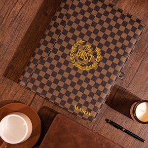 May include: A brown and black checkered leather folder with a gold embossed logo that says "The Best" and a laurel wreath. The folder has the name "MASON" printed on the bottom right corner.