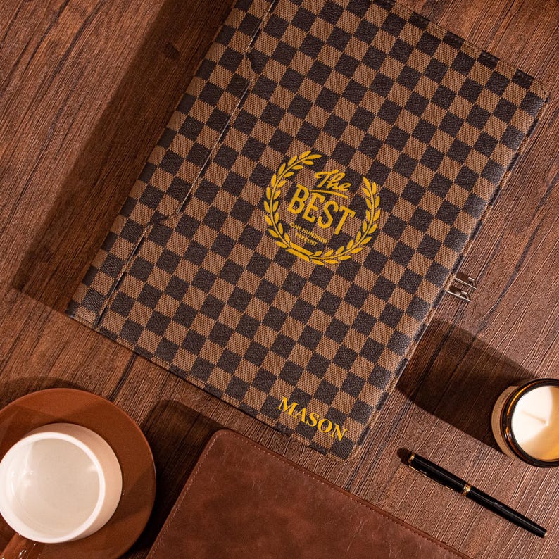 May include: A brown and black checkered leather folder with a gold embossed logo that says "The Best" and a laurel wreath. The folder has the name "MASON" printed on the bottom right corner.