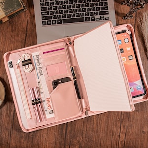 Może przedstawiać: Różowe skórzane portfolio z wieloma przegródkami, w tym na tablet, telefon, karty i długopisy. Otwarty, ukazujący notatnik i tablet. Widoczny napis "Mauris News" na wycinku z gazety. Całość na drewnianej powierzchni.