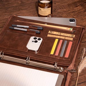 Pode incluir: Um fich&aacute;rio de couro marrom com fecho de z&iacute;per, com v&aacute;rios bolsos e slots para organiza&ccedil;&atilde;o. O fich&aacute;rio tem um mecanismo de argolas para segurar pap&eacute;is e inclui um bloco de notas, canetas e um smartphone.