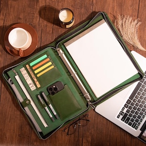 May include: A green leather zippered binder with a ring mechanism, a notepad, a phone pocket, and a zippered pocket. The binder is open and a laptop is partially visible.