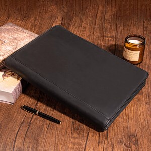 May include: A black leather zippered portfolio or folder with a smooth, textured surface. The portfolio is lying open on a wooden surface.