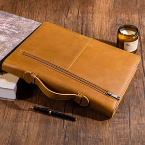 May include: A tan leather briefcase with a zip and a handle. A black pen lies on the wooden surface beside the briefcase. A candle in a glass jar and a book are also present.