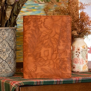 May include: A brown leather journal with an embossed floral design. The cover displays a detailed pattern of leaves and flowers. The journal has a zip closure and a diagonal seam. The background includes vases with dried flowers and a colourful painting.