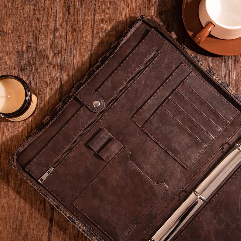 May include: Brown leather binder with multiple pockets and a zipper closure. The binder has a ring mechanism for holding papers.