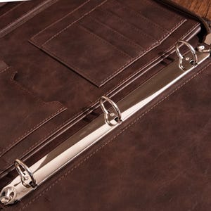 May include: A brown leather binder with three metal rings. The binder has multiple pockets and slots for holding papers and documents.