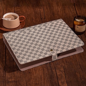 May include: A gray and white checkered pattern notebook or tablet cover with a snap closure. A cup of coffee with a spoon and a lit candle are also on the wooden surface.
