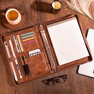 May include: A brown leather portfolio, zipped open to show a notepad, card slots, and a phone pocket. Two pens, a smartphone, and a few cards are visible. A cup of coffee and a candle are nearby.