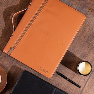 Może przedstawiać: Brązowe skórzane portfolio z zapięciem na zamek błyskawiczny i uchwytem. Słowo "MASON" jest wytłoczone w prawym dolnym rogu. Widoczny jest również czarny długopis i zapalona świeca w szklanym słoiku.