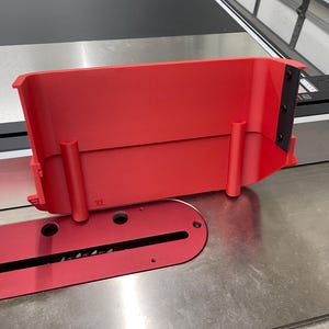 May include: A vibrant red table saw blade guard and throat plate. The guard is rectangular with two cylindrical supports. The throat plate features a blade slot and mounting holes. The saw is on a metal surface.