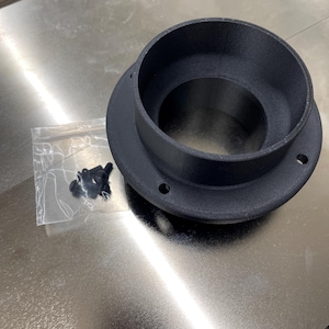 May include: A black, circular component with a wide base and a central opening. The component has several small holes around the base. A small plastic bag containing several small black fasteners is next to the component.
