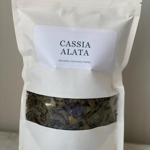 May include: A white pouch with a clear window displaying dried leaves. The pouch has a label that reads "CASSIA ALATA" above "Wholistic Harmony Herbs" in a simple, clean font. The leaves are a mix of green and brown.