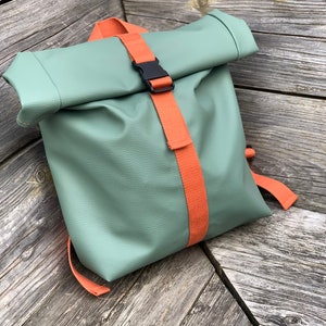 Op de afbeelding: Een mintgroene roll-top rugzak met oranje bandjes en een zwarte gespsluiting. De tas heeft een textuuroppervlak en verstelbare bandjes. De rugzak staat op een verweerde houten ondergrond.