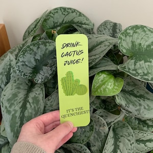 May include: A rectangular lime green bookmark featuring a dark green illustration of two cacti. The text DRINK CACTUS JUICE! is printed at the top, and the text IT'S THE QUENCHIEST! is printed at the bottom in a dark green sans-serif font.