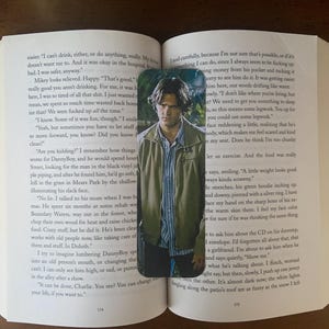 May include: A close-up of a bookmark featuring a man with dark hair and a green jacket. The bookmark is placed on an open book with text about a character named Danny Boy.