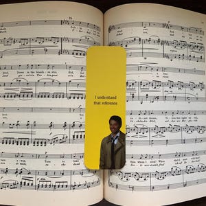 May include: A yellow bookmark with the text "I understand that reference" and an image of a man in a coat, placed in an open book of sheet music. The book is open to a page of musical notation.