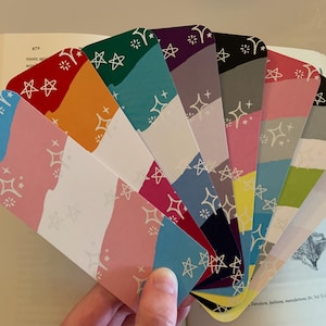 May include: A collection of colorful bookmarks fanned out, each with a unique design. The bookmarks feature a mix of colors like red, orange, blue, pink, and green, with a white starburst pattern. They are held in front of an open book.