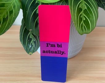 Peut inclure: Un autocollant rectangulaire aux couleurs rose, violet et bleu. Le texte "I'm bi actually" est imprimé sur l'autocollant. L'autocollant est placé devant une plante verte avec des feuilles rayées blanches et vertes.
