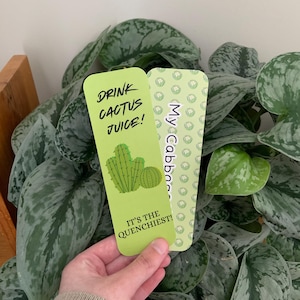 Peut inclure: Deux marque-pages rectangulaires aux coins arrondis. L'un est vert clair avec une illustration de cactus et le texte DRINK CACTUS JUICE! IT'S THE QUENCHIEST! L'autre est vert plus pâle avec un motif de choux répété et le texte My Cabbages.
