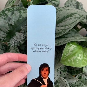 Op de afbeelding: Een lichtblauwe rechthoekige boekenlegger met de gedrukte tekst "Hey girl, are you improving your mind by extensive reading?" boven een fotografische afbeelding van een persoon met donker haar in een zwarte jas en een wit overhemd.